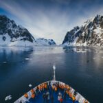 Cruise ship bow with passengers looking out over dark blue ocean with scattered icebergs towards cliffs of Fjord coastline covered in snow. Cruise Antarctica Travel Antarctica