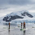 Four SUP'ers paddle among small icebergs in front of a glacier ice wall and snow covered mountain background. Antarctic expedition cruise Polar cruise
