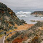 Passengers walk down wooden trail towards small bay with icebergs in sight. Arctic travel Polar region travel Greenland cruise