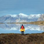 A lone hiker stares across the mirror waters to the snow capped mountain ranges in the distance. Greenland hiking in the Arctic region. Polar travel Greenland cruise