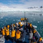Passengers standing on bow of cruise ship viewing the Arctic landscape of ice spotted water and glacier ice wall and snow covered mountain range. Arctic cruise Arctic travel