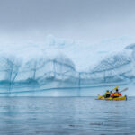 Two paddlers paddling up to a large white with blue ice lines iceberg. Travel Antarctica Holiday in Antarcitca