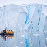 A zodiac boat with passengers cruises in front of paper white ice wall above mirror like water. Dark blue crevasse lines can be seen in the ice wall. Antarctica cruise Polar travel