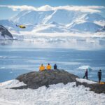 Passengers sit a top a rocky outcrop above snow viewing a bay of the ocean before a glacier wall and mountain range as a expedition helicopter flies above the flat blue ocean. Antarctica vacation Polar holiday