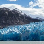 Blue of glacier wall which jagged ice peaks stands in contrast to sea water and brown mountain ranges in distance. Chile Glacier travel. Chile Cruise