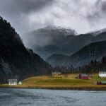 Landscape of Mostraumen, - Norway with water before grassland with small houses against backdrop of forested mountain range with ice capped peaks. Cruise in the North Sea