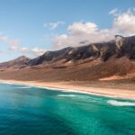 Stunning coastline of Playa de Cofete Cape Verde Island with blue waters breaking on sandy shore which rises to steep mountain range. Sea cruise Cape Verde