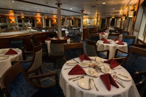 Dinning room of the Ocean Adventurer polar ship. Many round tables spaciously space throughout the room with wooden material covered dinning chairs. Table laid out in simplistic classy way.