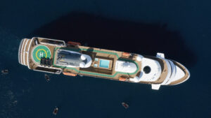 A birds eye view of the ship World Explorer in deep dark blue ocean waters. The stern on the left of the picture has a helicopter landing pad where zodiac boats are stored. The middle of the ship on the top deck has an outside swimming pool. Towards the right of the picture is the stern of the ship, from this view one can see the ample wooden deck viewing decks that passengers can enjoy whilst aboard and in the Arctic and Antarctic regions.