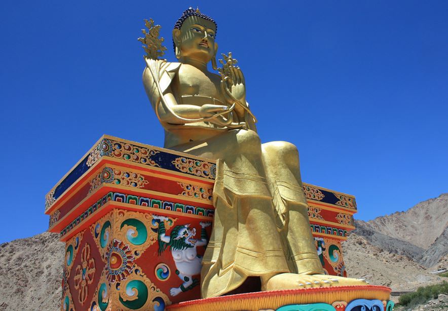 Large gold Buddha statue on top colorful patterned pedestal in India against the blue of the sky behind it. Small group tour