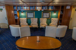 Library on board the Ocean Adventurer. Classy comfortable cream single chairs with wooden tables setup with wall booked shelf behind. A great place to read up on the adventures of the Arctic and Antarctic regions passengers are travelling through.