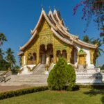 Buddha temple with golden detailed outside wall with white steps leading up set amount green grass and tall palm tree garden.| Itinerary stop on Asian river cruise along Meekong river.