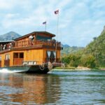 River cruise ship in Asia cruising along the river with drastic mountain background littered with green foliage. The boat is a stunning wood and classic shape and one can view the cabin windows.