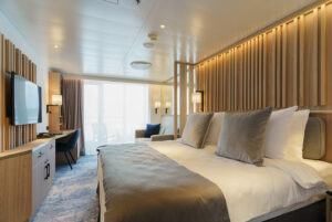 A balcony suite cabin with double bed configuration aboard the Ultramarine ship - designed for Arctic and Antarctic trips. Sleekly designed with wooden finished with grey and white bedding. In the background, the window doors lead to outside balcony.