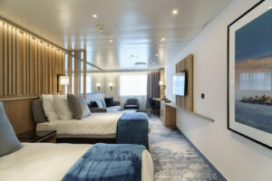 An Explorer Suite cabin on board the Ultramarine ship designed for polar travel. Classy wooden finishes complete the blue and grey bedding of the twin single beds in the foreground. In the background the living area sits in front of a large exterior window.