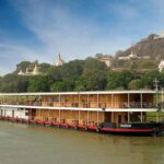 River cruise ship Kindat Pandaw on Brahmaputra River India. Class 2 deck wooden ship moored on bank of river with shrines in the background atop a hill.