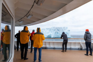 Passengers enjoying the view of a massive white iceberg from the outside viewing deck the ship in the polar regions.