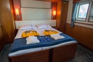 Twin window suite cabin of the ship Ocean Adventurer in a double bed setup. Simple and sleek wooden finishes compliment the white and blue bed fittings. Polar jackets are laid out on the beds.