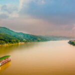 River cruise on the Red River of Vietnam. A view of the ship from afar in the big river of reddish tinged water. Banks of the river are green with rolling hills. River cruise travel.
