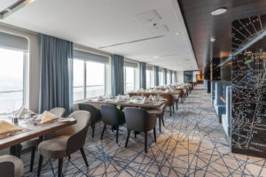 Dining hall onboard the Ultramarine ship. Modern design carpeted flooring with blue backed grey interior dinning chars at wooden tables next to large windows. A stylish way to enjoy meals whilst exploring the Arctic and Antarctic regions.