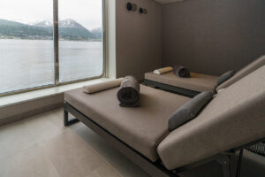 Comfortable grey massage tables of the Spa and Sauna aboard the polar expedition ship Ultramarine. The floor to ceiling window shows the passing scenery of the Arctic ocean and ice capped mountains.