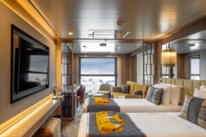 An Infinity Suite Cabin aboard the ship World Explorer, designed for Polar travel. Two twin beds facing a flat screen TV in the foreground with a couch and coffee table and chairs in the background next to the floor to ceiling window of the outside where one can see ice covered mountains in the distance. A sleek wooden finish portrays the comfort of the cabin.