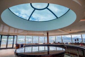 A view of the Observation Lounge of the ship World Explorer designed for Polar Travel. A large sunlight centers the roof in the picture with floor to ceiling widows as the walls creating a panoramic view of the outside.