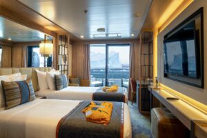 View of Veranda Suite cabin aboard the ship World Explorer. Two twin beds with the yellow expedition jackets on tp face a large TV screen. Further back the floor to ceiling window door opens onto the balcony where the passing view is of a massive iceberg in the ocean. Polar travel